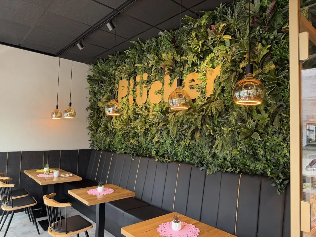 Sitzbereich der Bäckerei Plücker in Wabern mit Pflanzenwand, Polsterbank und Holztischen.
