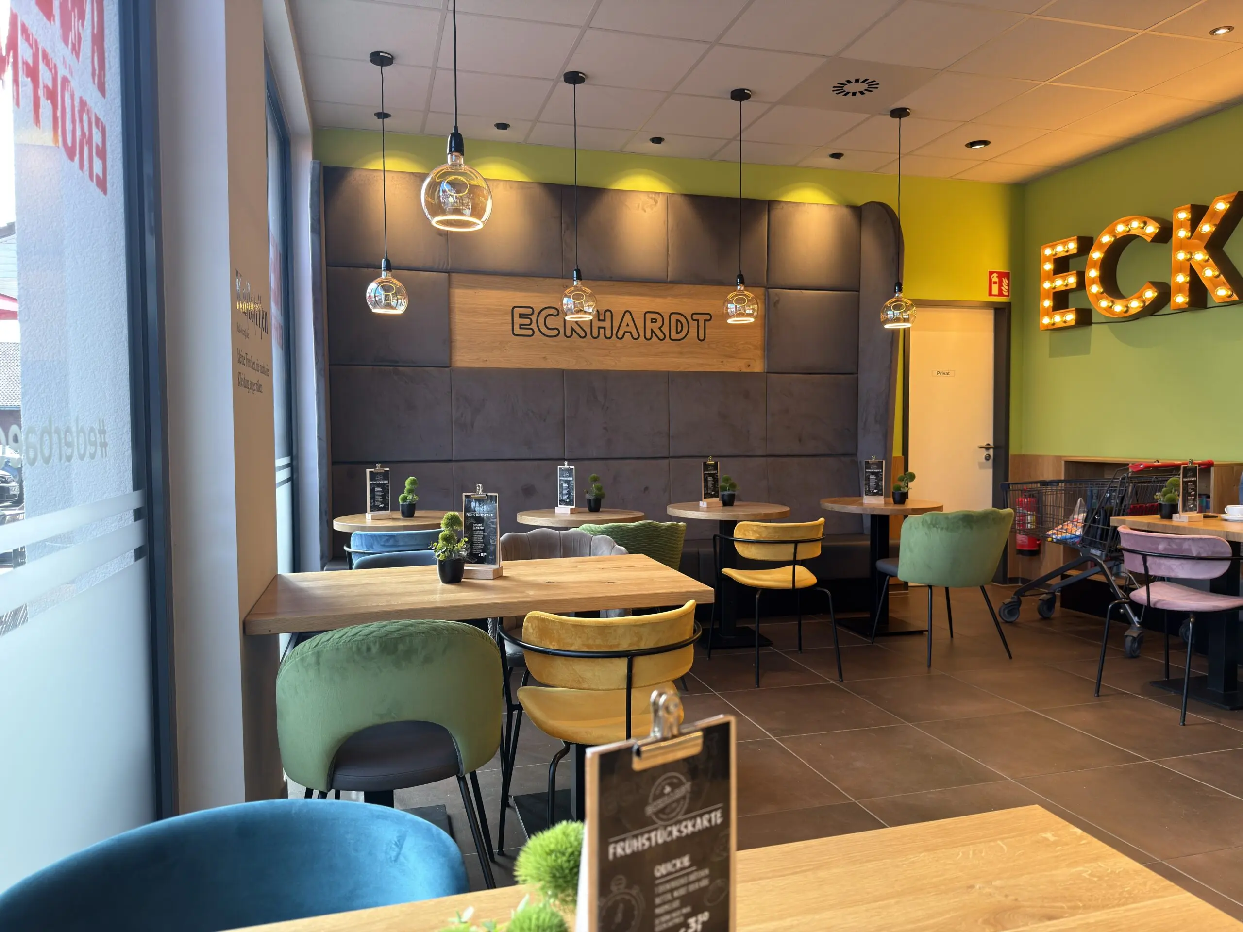 Sitzbereich der Bäckerei Eckhardt mit farbigen Stühlen, Holztischen, Pendelleuchten und gepolsterter Wand mit Logo.