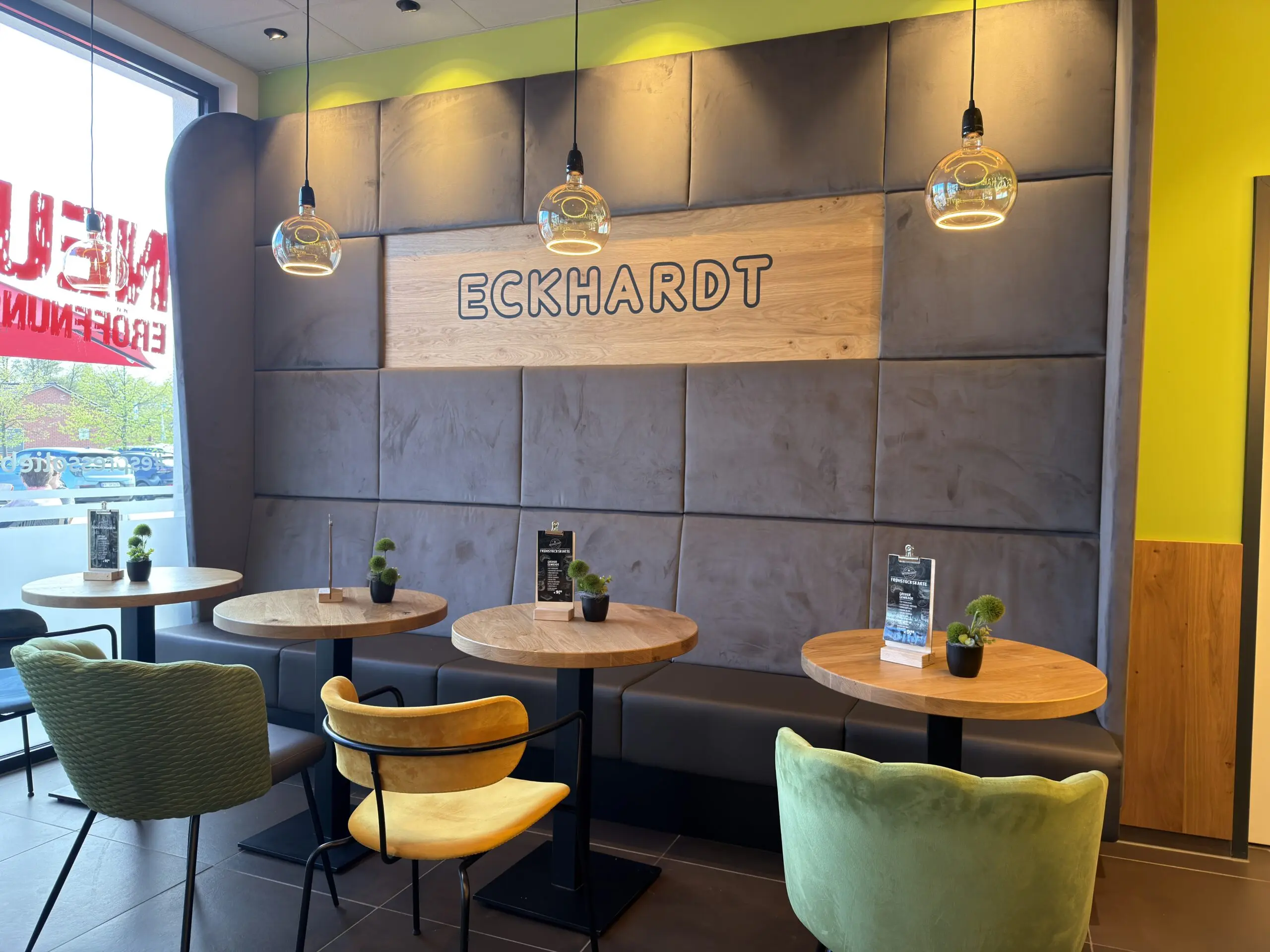 Sitzbereich der Bäckerei Eckhardt mit farbigen Stühlen, runden Holztischen und gepolsterter Wand mit Logo.