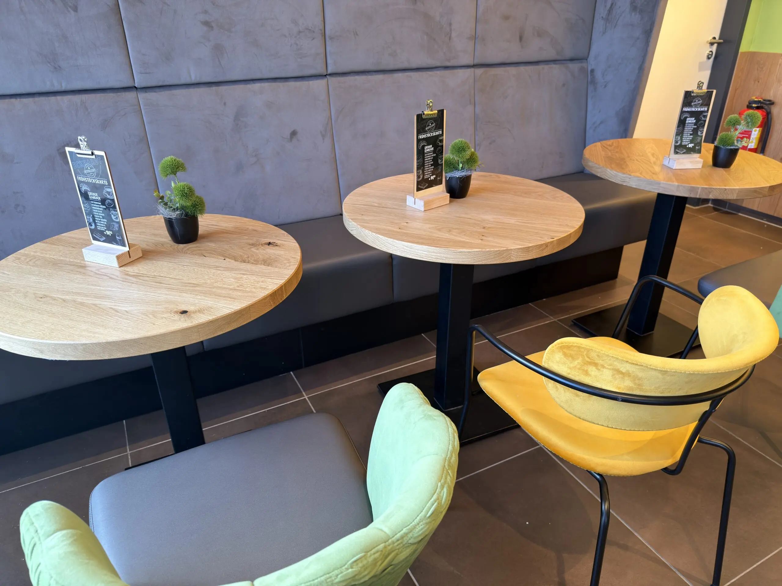 Detailaufnahme des Sitzbereichs der Bäckerei Eckhardt mit runden Holztischen, farbigen Stühlen und gepolsterter Wand.