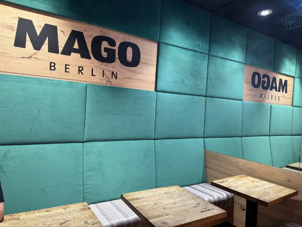 Sitzbereich von MAGO im Märkischen Zentrum Berlin mit türkis gepolsterter Wand, Holzflächen und Markenschriftzug.