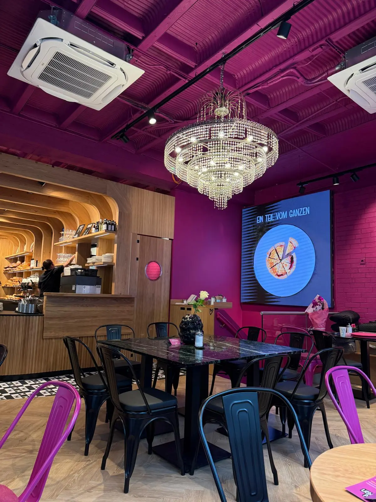 Sitzbereich der Bäckerei Huck in Frankfurt mit Kronleuchter, pinker Wandgestaltung, Holztischen und gemütlichen Sitzplätzen.