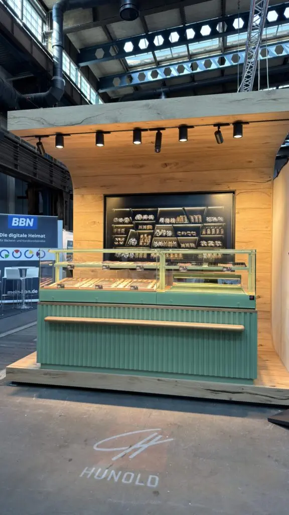 Pop-up-Messestand von Ladenbau Hunold auf dem inpraxi Handelstag mit grüner Bäckereitheke und projizierter Hunold-Logo-Beleuchtung auf dem Boden.